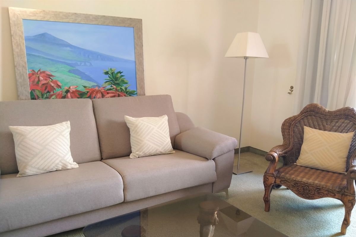 Divano beige con cuscini, sedia intrecciata e lampada, quadro di Tenerife sullo sfondo.