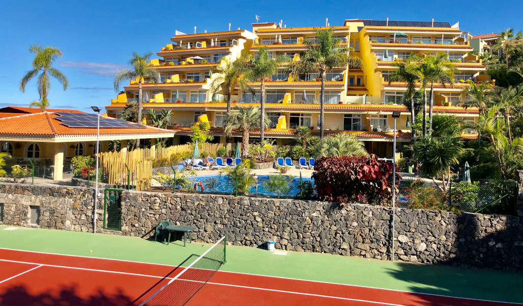Edificio giallo con piscina e palme a Tenerife, vista da un campo da tennis.