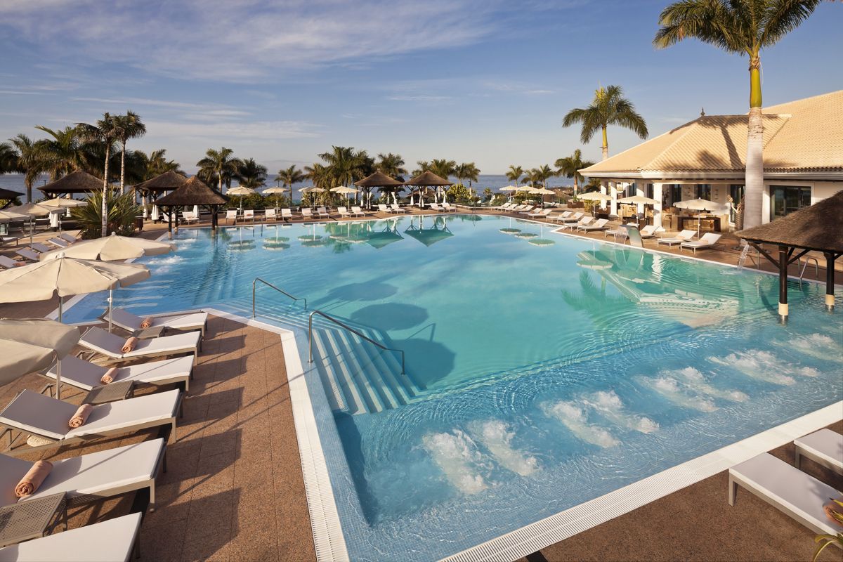 Piscina con lettini e palme nell'hotel a Tenerife, cielo sereno e mare sullo sfondo.