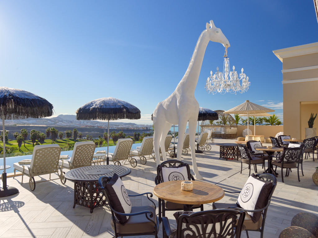 Terrazza con tavoli, sedie, una giraffa bianca statua e vista su Costa Adeje, Tenerife.