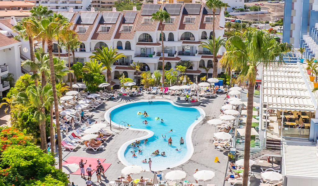 Piscina affollata circondata da lettini e palme in un hotel a Playa de las Américas.