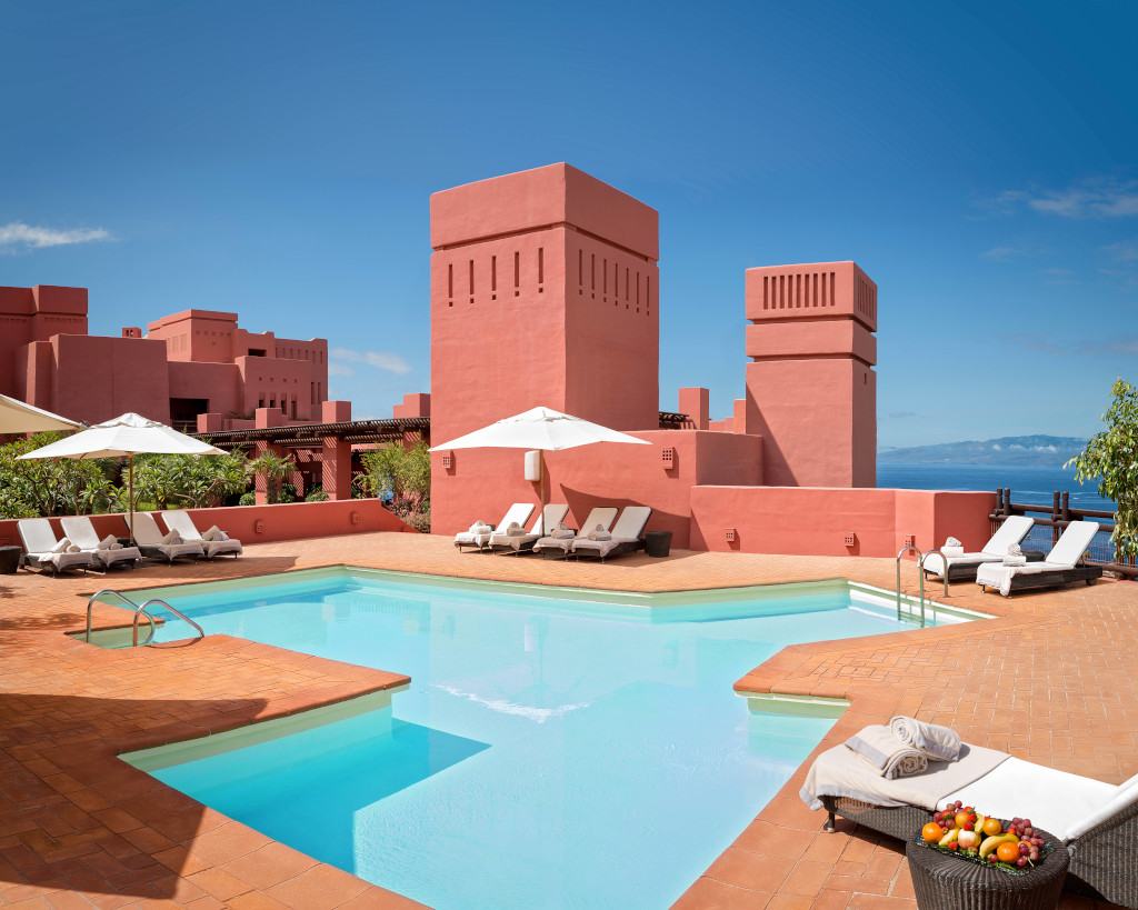 Piscina con lettini bianchi, edifici rossi e vista sull'oceano a Tenerife.