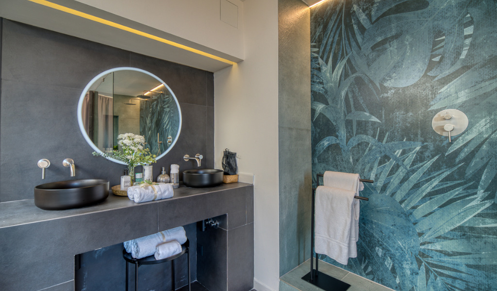 Bagno moderno con specchio rotondo, lavandini neri e parete con foglie a Tenerife.