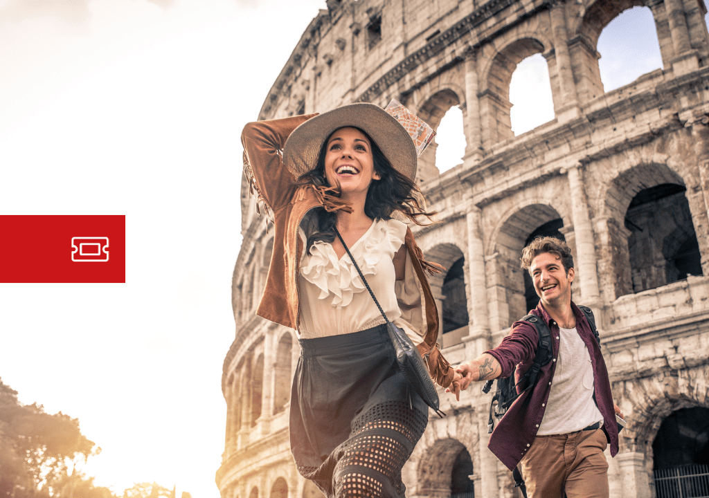 Coppia di turisti sorridenti che corre davanti al Colosseo di Roma al tramonto, con zaini e abbigliamento casual.