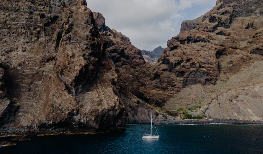 Barca a vela sulla costa rocciosa di Tenerife con acque azzurre e montagne sullo sfondo.