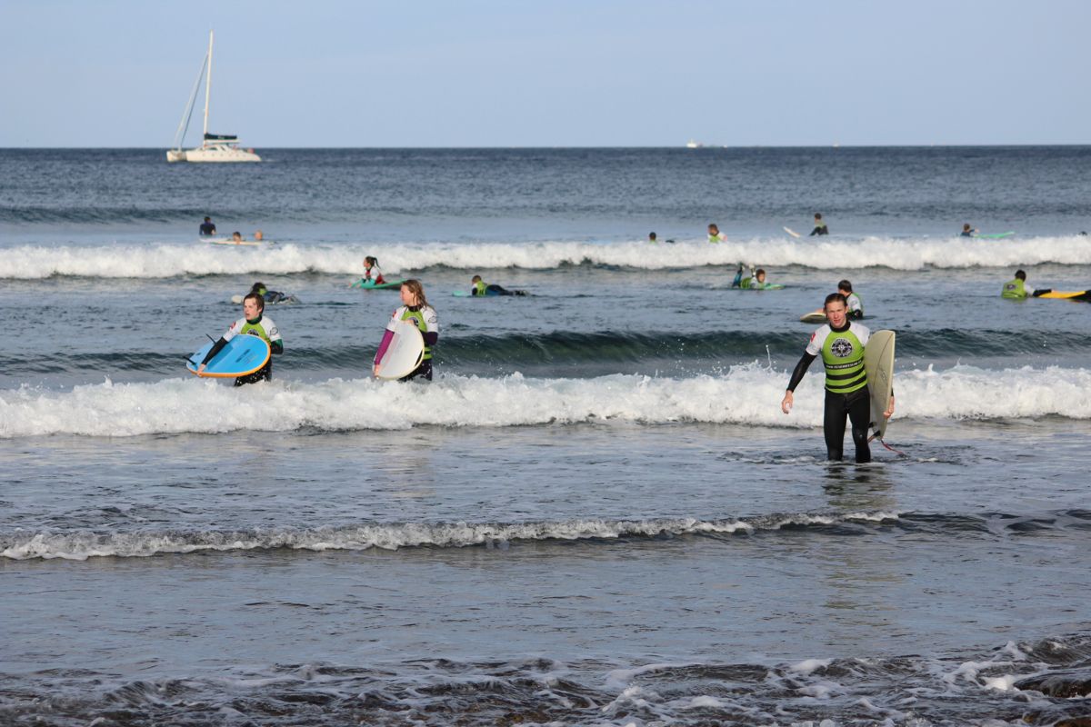 Surfisti con tavole a Playa de Las Américas, Tenerife, con una barca a vela sullo sfondo.