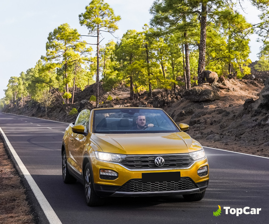 Auto gialla sulla strada circondata da pini a Tenerife. Logo TopCar.