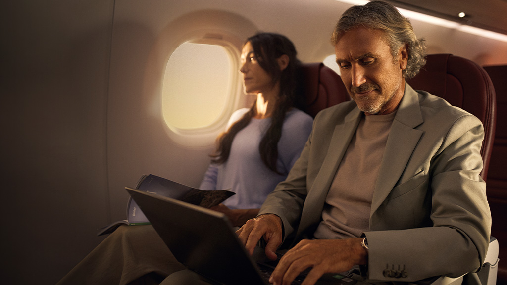 Persona che usa il portatile su un aereo, accompagnata da un passeggero che legge.