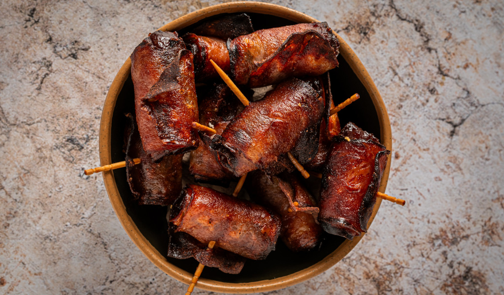 Salsicce avvolte nel bacon con bacchette su sfondo di pietra.