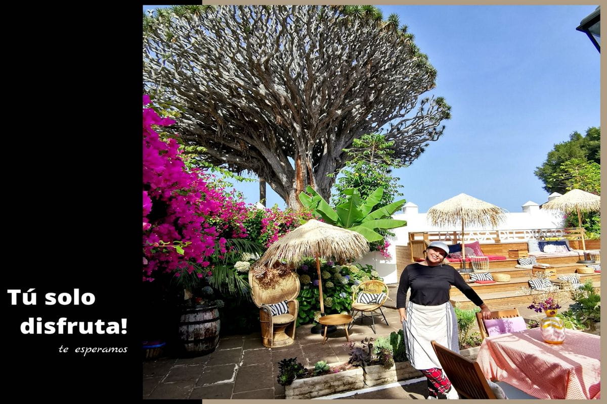 Patio con vegetazione, ombrelloni di paglia e una persona a Tenerife. Testo: Divertiti e basta! Ti stiamo aspettando.