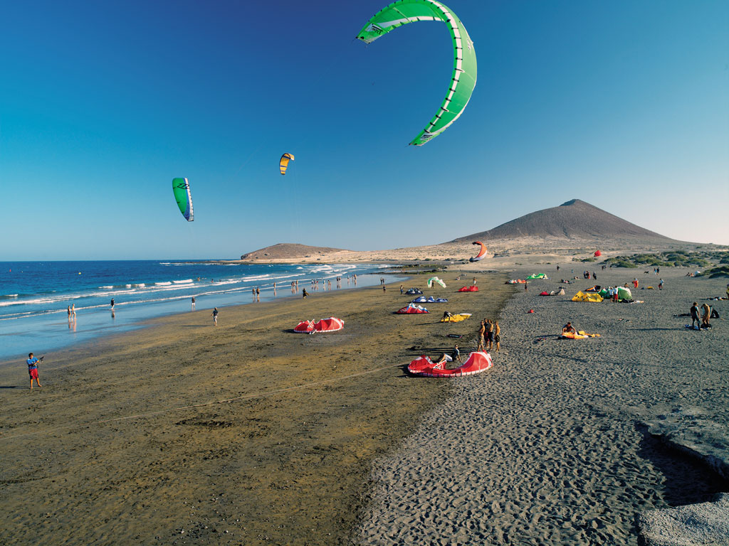 Persone che fanno kitesurf sulla spiaggia di El Médano, Tenerife, con montagne sullo sfondo.
