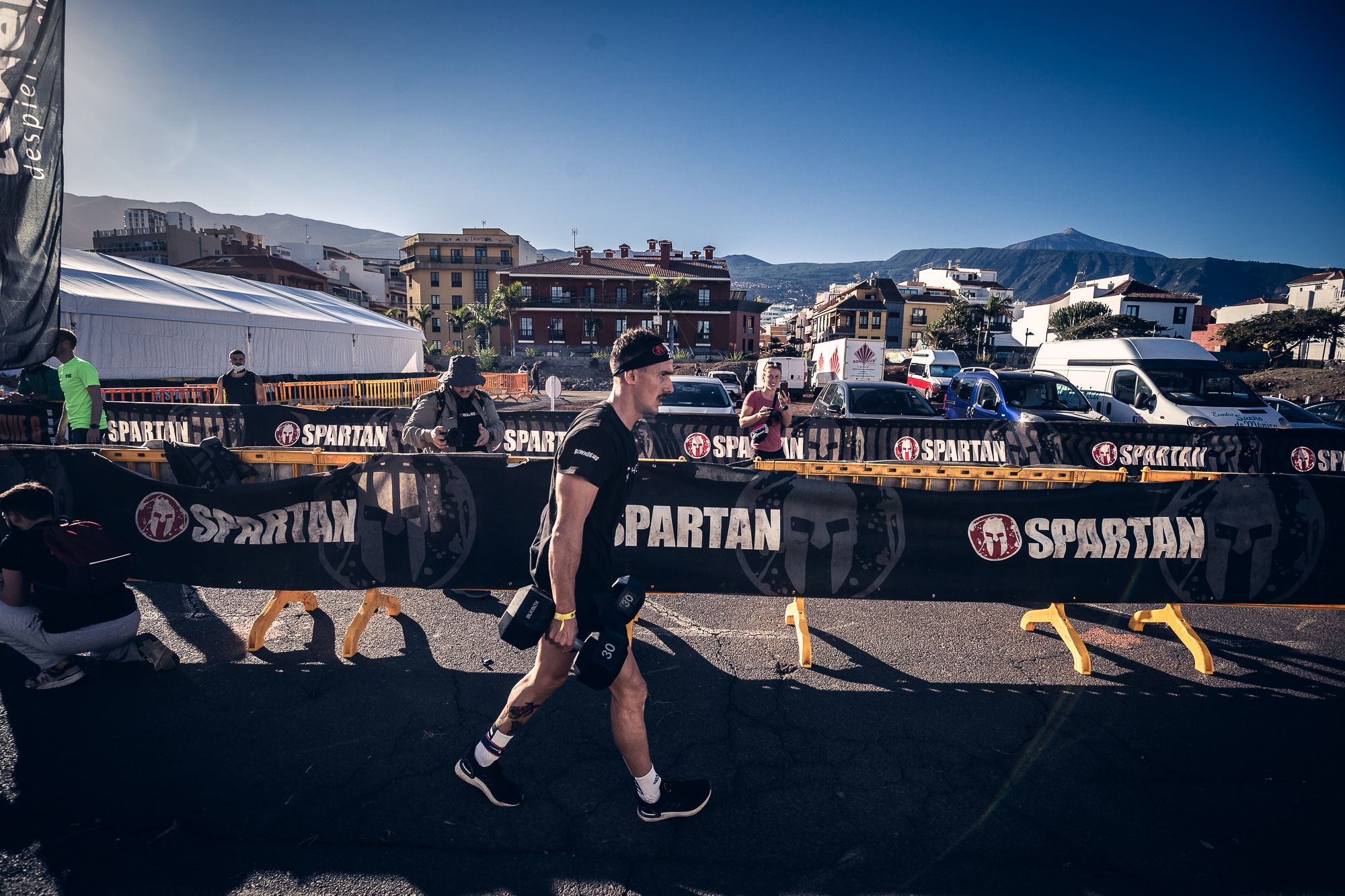 Concorrente nell'evento Spartan a Tenerife portando pesi, edifici e montagne sullo sfondo.