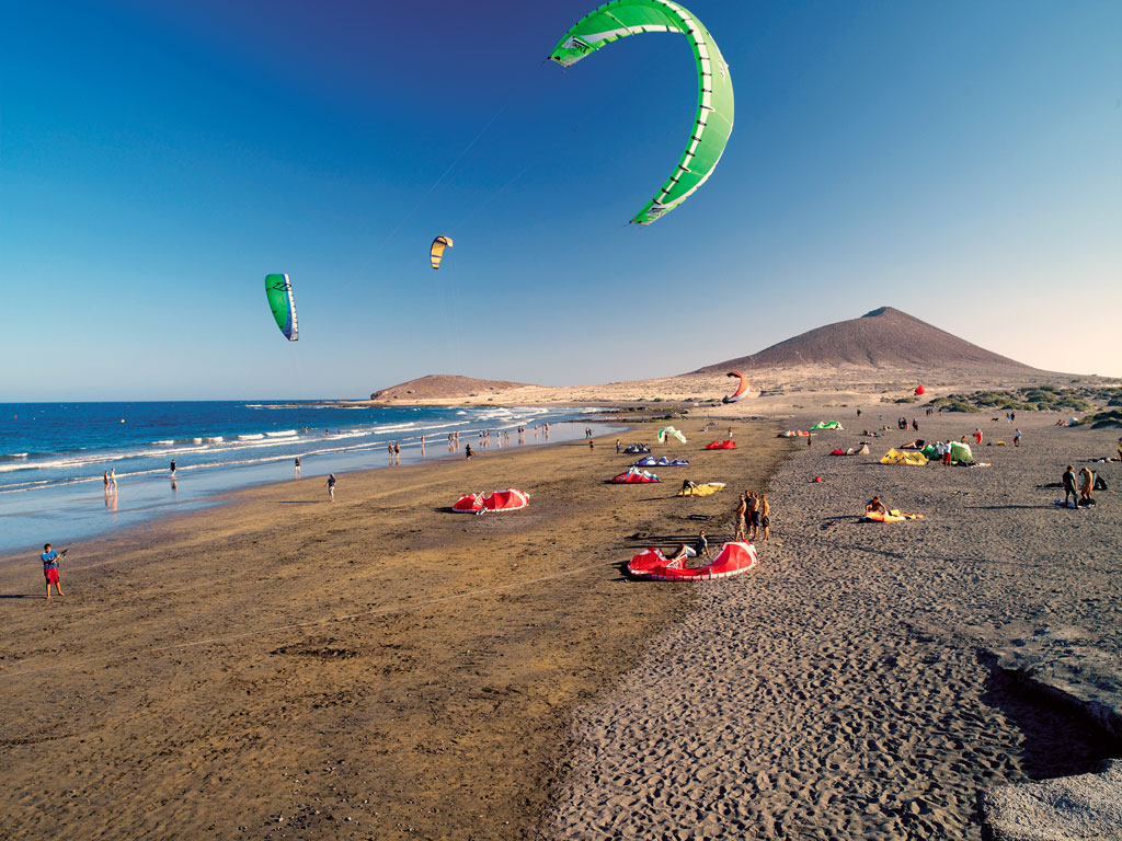 Persone che fanno kitesurf sulla spiaggia di El Médano, Tenerife, con montagne sullo sfondo.