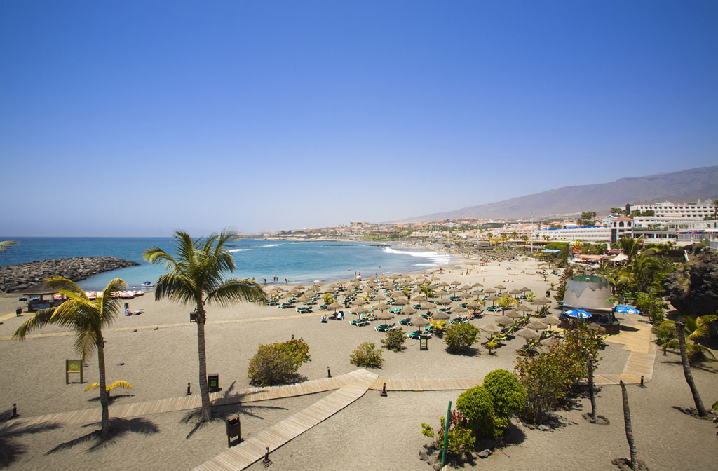 Spiaggia sabbiosa con palme e sdrai a Tenerife, vista sul mare e sugli edifici sullo sfondo.