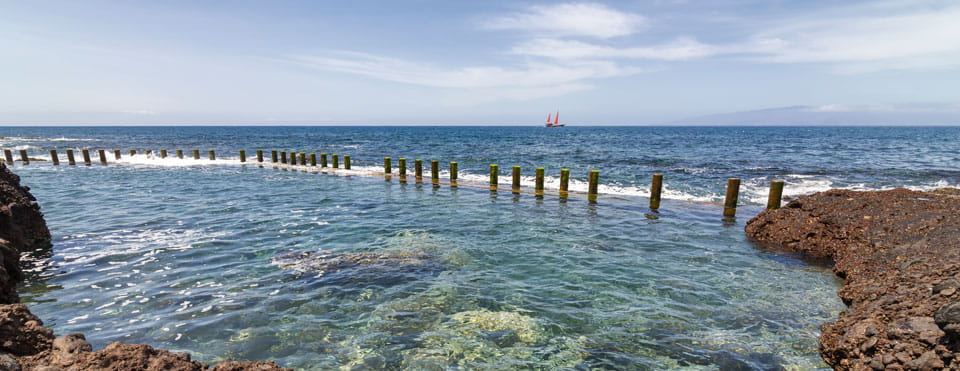 Acqua cristallina e pali di legno sulla costa di Tenerife, barca con vele rosse sullo sfondo.